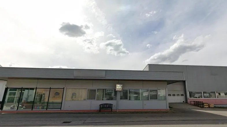Ansicht eines Gewerbegebäudes mit einem vorderen Bürobereich und einer dahinterliegenden Lagerhalle unter bewölktem Himmel.