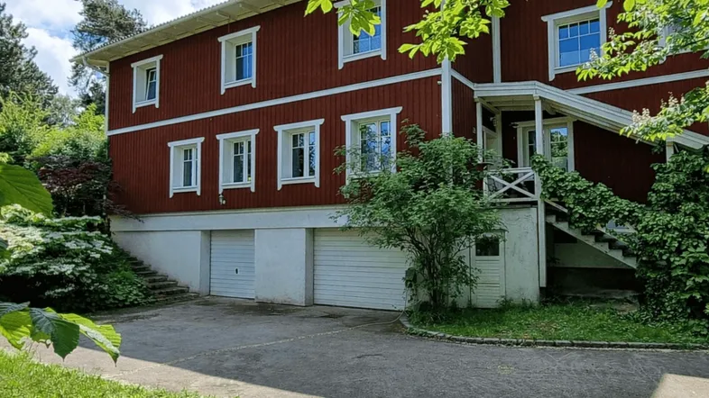 Rotes Holzhaus mit weißen Fenstern und Garagen im Untergeschoss, umgeben von viel Grün.
