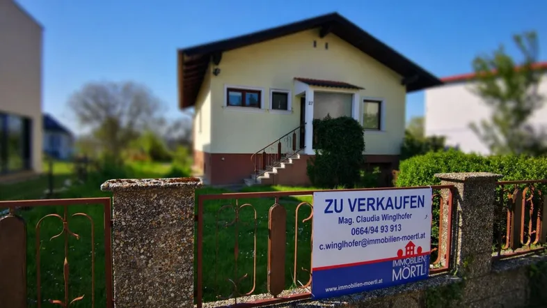Einfamilienhaus mit Vorgarten und Verkaufsplakat in ländlicher Umgebung.