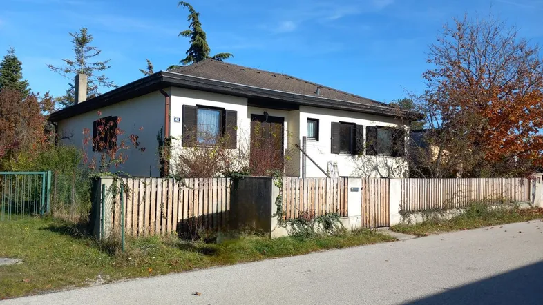 Einfamilienhaus mit kleiner Umzäunung und Vorgarten, umgeben von Bäumen unter blauem Himmel.