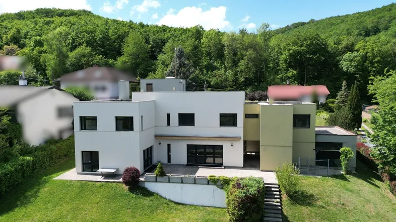 Moderne Villa mit Flachdach und weitläufigem Garten in hügeliger Landschaft, umgeben von Bäumen.
