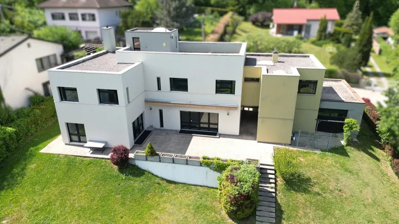 Moderne Villa mit Flachdach, heller Fassade und großem Gartenbereich in grüner Umgebung.