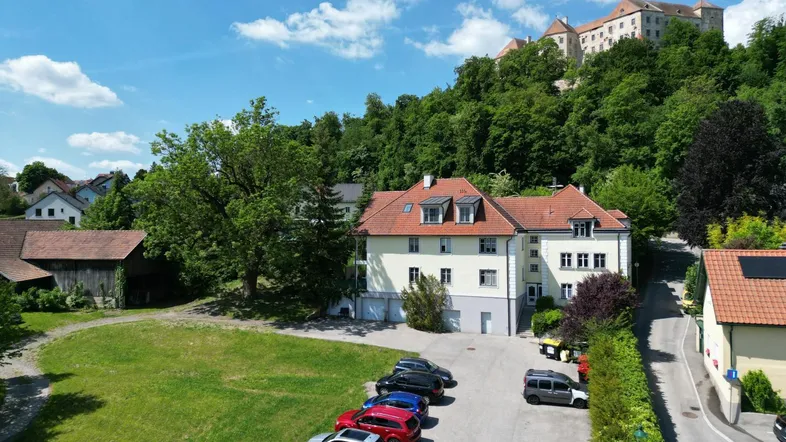 Luftaufnahme des Gebäudes mit Parkplätzen, umgeben von Grünflächen und einem Schloss im Hintergrund.