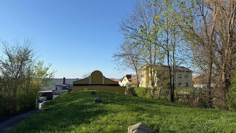 Grüne Hügellandschaft mit Bäumen und Blick auf entfernte Gebäude unter blauem Himmel.