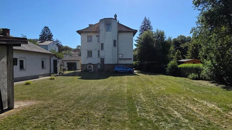Großes Grundstück mit Rasenfläche und einem älteren Wohnhaus unter blauem Himmel.