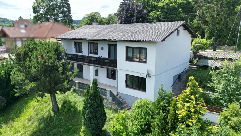Freistehendes Einfamilienhaus mit weißer Fassade und Satteldach, umgeben von viel Grün und Bäumen.