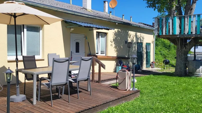 Große Holzterrasse mit Esstisch und Sonnenschirm, umgeben von grünem Rasen und einem Baumhaus.
