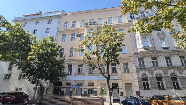 Ansicht der historischen Fassade eines Mehrfamilienhauses mit Bäumen und parkenden Autos auf der Straße.