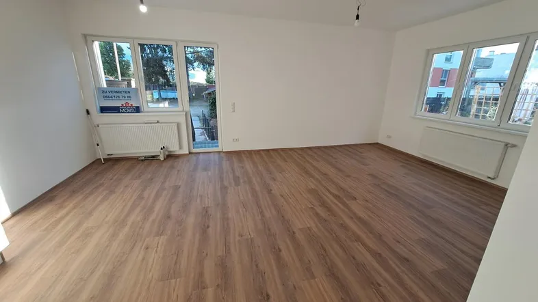 Helles Wohnzimmer mit Laminatboden, großen Fenstern und Zugang zu einem Außenbereich.
