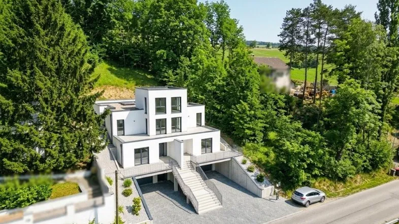 Moderne weiße Villa mit mehreren Ebenen, umgeben von üppigem Grün und einer Zufahrtsstraße.
