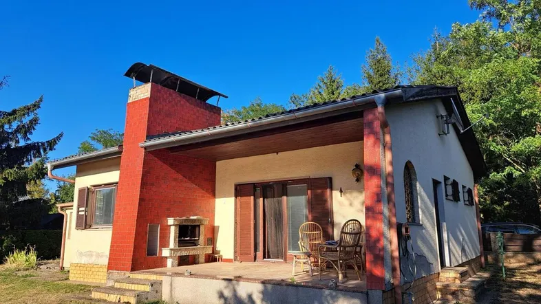 Hausfassade mit überdachter Terrasse, Außenkamin und Sitzgelegenheiten im Freien unter blauem Himmel.