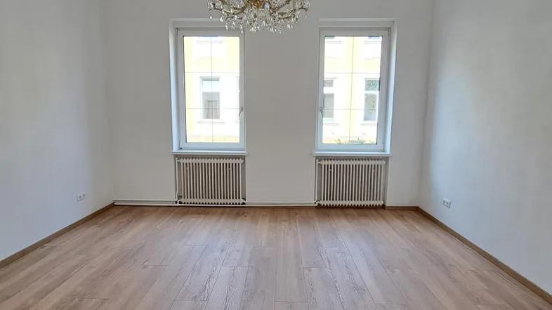 Heller, leerer Raum mit Holzboden, zwei Fenstern und einem eleganten Kronleuchter.