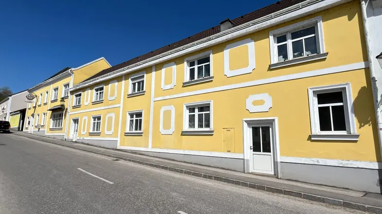 Gelbes Gebäude mit weißen Fensterrahmen und traditioneller Architektur an einer leicht ansteigenden Straße.