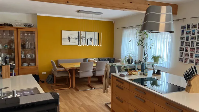 Offener Wohn- und Essbereich mit moderner Küche, Holzbalken und gelber Akzentwand.
