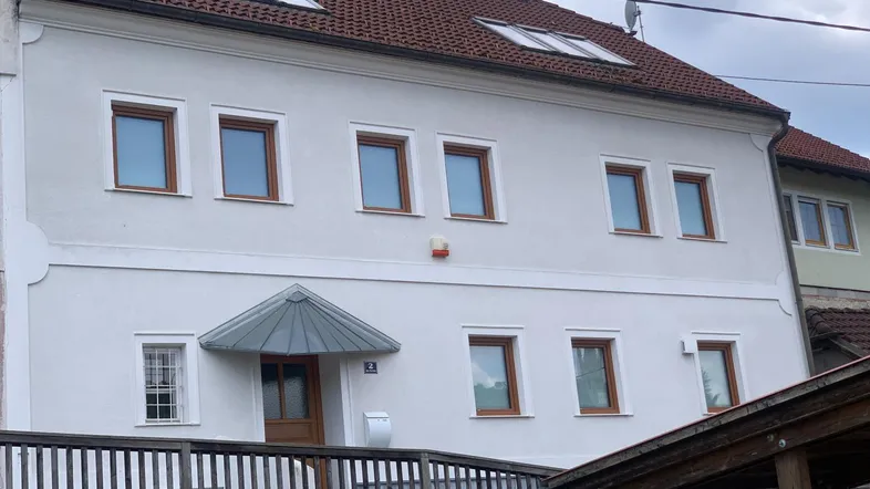 Weiß verputztes Gebäude mit braunen Fenstern und rotem Ziegeldach unter bewölktem Himmel.