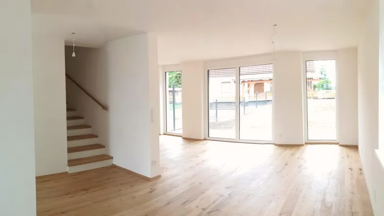 Heller, leerer Wohnbereich mit Holzboden, großen Fenstern und Treppenaufgang zu den oberen Etagen.