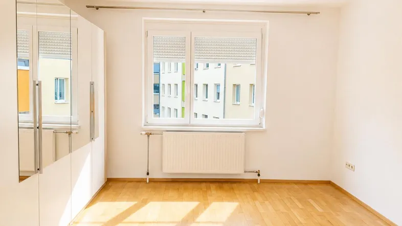 Helles Schlafzimmer mit Parkettboden, großem Fenster und weißem Spiegelschrank.