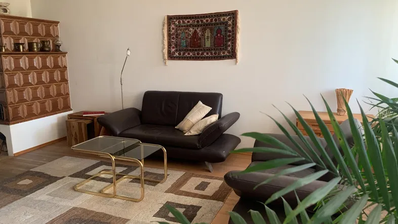 Gemütliches Wohnzimmer mit Kachelofen, dunklen Ledersofas und einem Teppich mit geometrischem Muster.