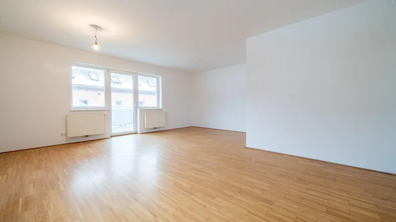 Helles Wohnzimmer mit Parkettboden, weißen Wänden und Zugang zu einem Balkon.