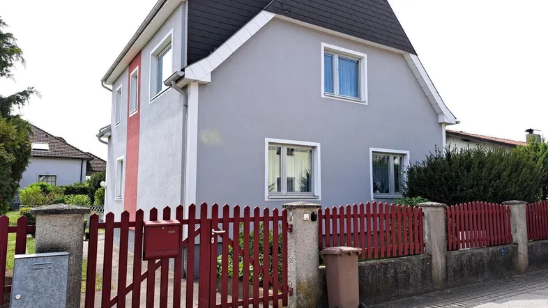 Einfamilienhaus mit grauer Fassade, rotem Zaun und klassischem Satteldach.