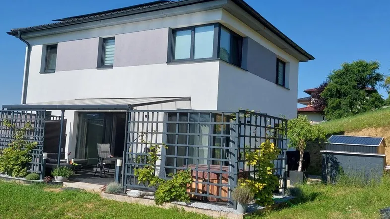 Modernes Haus mit überdachter Terrasse, Whirlpool und Gartenbereich unter blauem Himmel.