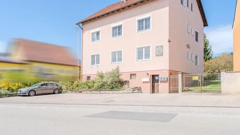 Mehrstöckiges Wohngebäude mit hellrosa Fassade, braunem Dach und weißen Fenstern an einem sonnigen Tag.