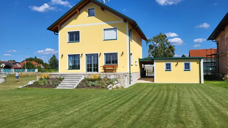 Gelbes Einfamilienhaus mit gepflegtem Rasen, Terrasse und Carport unter blauem Himmel.