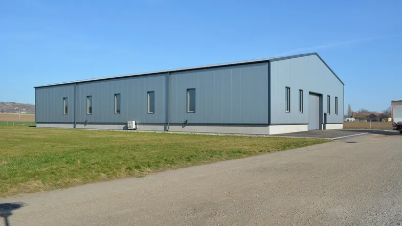 Langes, modernes Industriegebäude mit blaugrauer Metallfassade und kleinen Fenstern auf einer grünen Wiese.