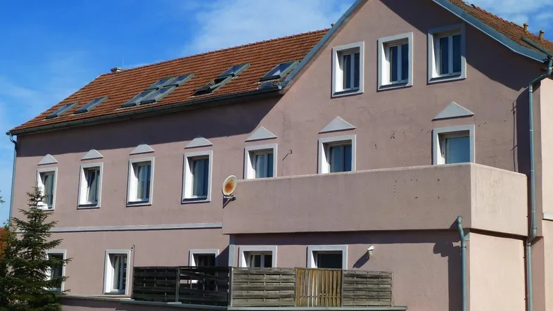 Ansicht eines Wohngebäudes mit rosa Fassade, Satteldach und einem Balkon im ersten Stock.