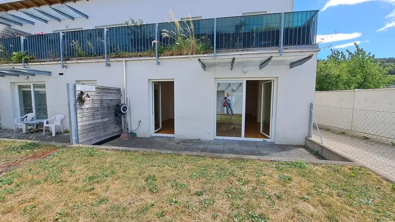 Außenansicht eines modernen Gebäudes mit Terrasse, kleinem Gartenbereich und Balkon im Obergeschoss.