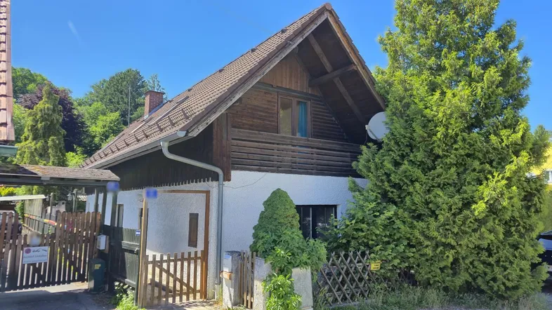Traditionelles Haus mit Holzfassade und Satteldach, umgeben von grünen Bäumen unter blauem Himmel.