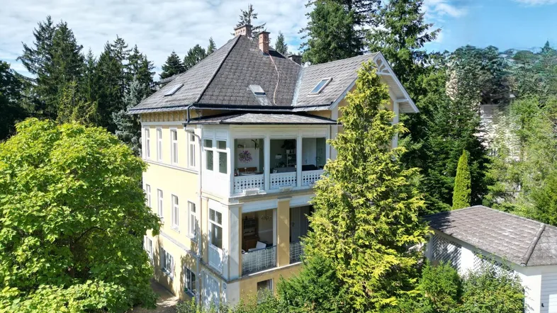 Historische Villa mit gelber Fassade, mehreren Balkonen und umgeben von altem Baumbestand.
