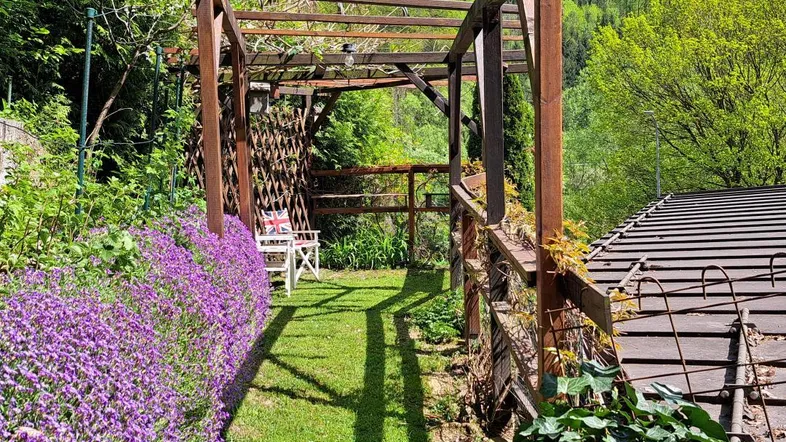 Grüner Gartenweg mit blühenden Pflanzen, Holzpergola und Blick auf bewaldete Hügel.