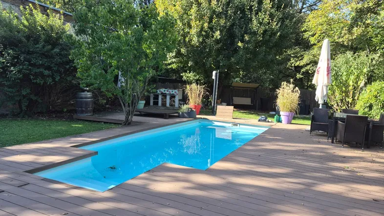 Großer Gartenbereich mit eingelassenem Pool, Holzterrasse und Sitzgelegenheiten unter Bäumen.