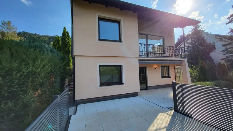 Moderne Hausfassade mit sonniger Terrasse und Balkon, umgeben von grüner Landschaft.