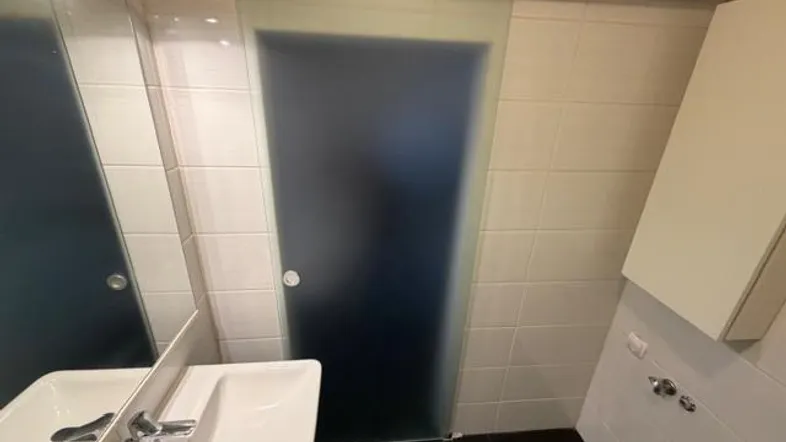 Modernes Badezimmer mit Waschbecken, Spiegel und einer Milchglas-Schiebetür, hell gefliest.