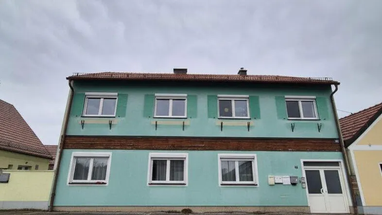 Fassade eines Wohnhauses mit mintgrüner Farbe und grünen Fensterläden unter bewölktem Himmel.