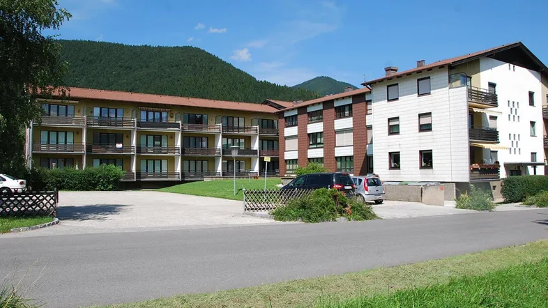 Mehrfamilienhaus mit mehreren Balkonen und Parkplätzen, umgeben von grüner Landschaft und Bergen.