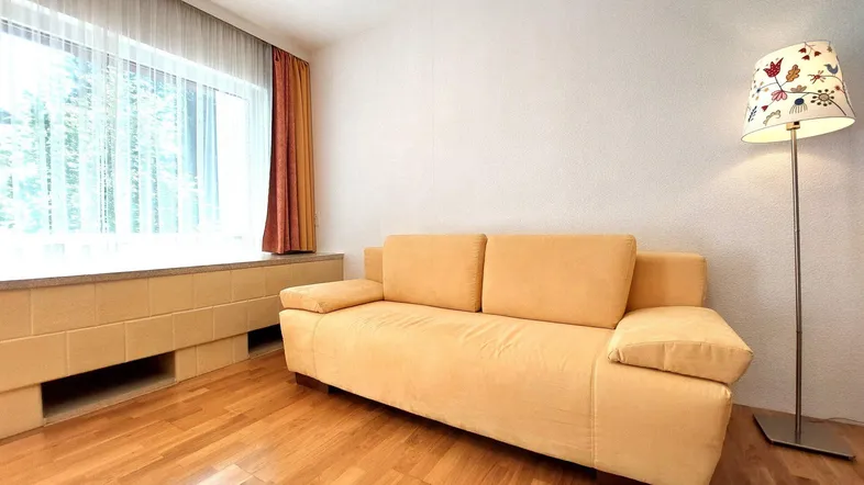 Helles Wohnzimmer mit beigem Sofa, Parkettboden und großem Fenster mit Vorhängen.