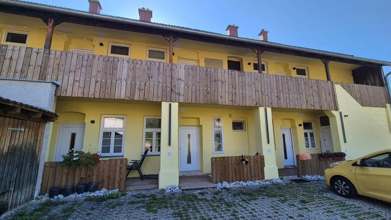 Gelbe Fassade eines Mehrfamilienhauses mit Holzbalkon und gepflastertem Vorplatz.