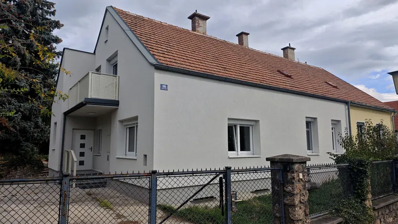 Vorderansicht eines renovierten Einfamilienhauses mit Balkon und Satteldach unter bewölktem Himmel.