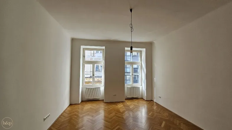 Helles Zimmer mit Fischgrätparkett und zwei großen Fenstern, das viel Tageslicht hereinlässt.