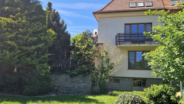 Hausfassade mit Balkon und Gartenbereich unter blauem Himmel, umgeben von Bäumen.