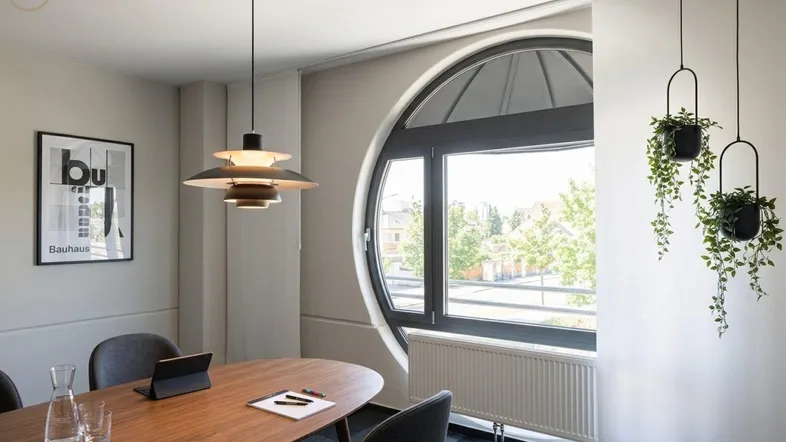 Moderner Besprechungsraum mit ovalem Holztisch, Designerlampe und großem Rundfenster mit Ausblick.