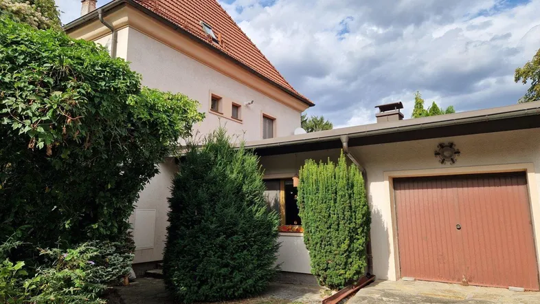 Hausfassade mit rotem Ziegeldach, angrenzender Garage und gepflegtem Vorgarten unter bewölktem Himmel.