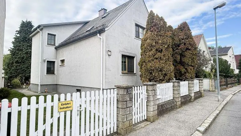 Weißes Einfamilienhaus mit Satteldach, Vorgarten und weißem Lattenzaun an der Straße.