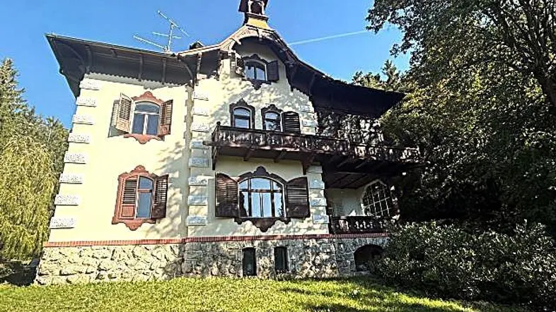 Historische Villa mit markanter Architektur, Holzbalkonen und Steinsockel, umgeben von Bäumen.