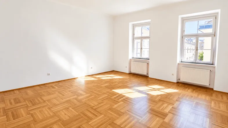 Helles, leeres Zimmer mit Fischgrätparkettboden und zwei großen Fenstern, die viel Tageslicht hereinlassen.