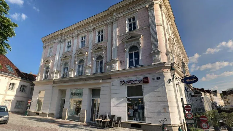 Historisches Gebäude mit aufwendiger Fassade und Geschäften im Erdgeschoss an einer gepflasterten Straße.