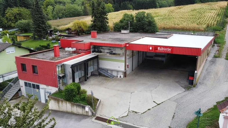 Großes Gewerbegebäude mit roter Fassade und weitläufigem Vorplatz, umgeben von grüner Landschaft und Feldern.